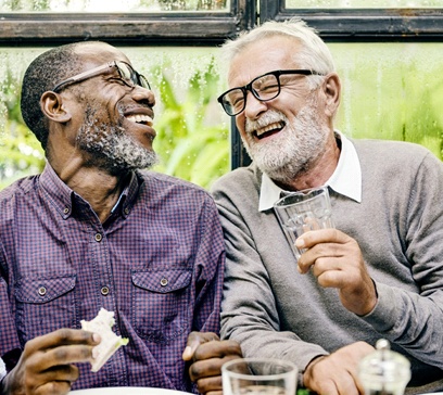 Two older men laughing and enjoying a meal while embracing the ability to eat confidently with their dental implants in Bethel Park