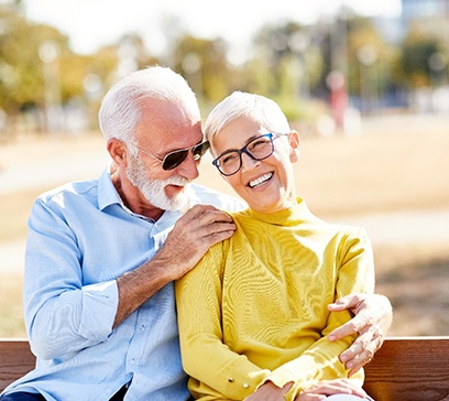 An older man and woman sitting on a bench and smiling after receiving dental implants in Bethel Park